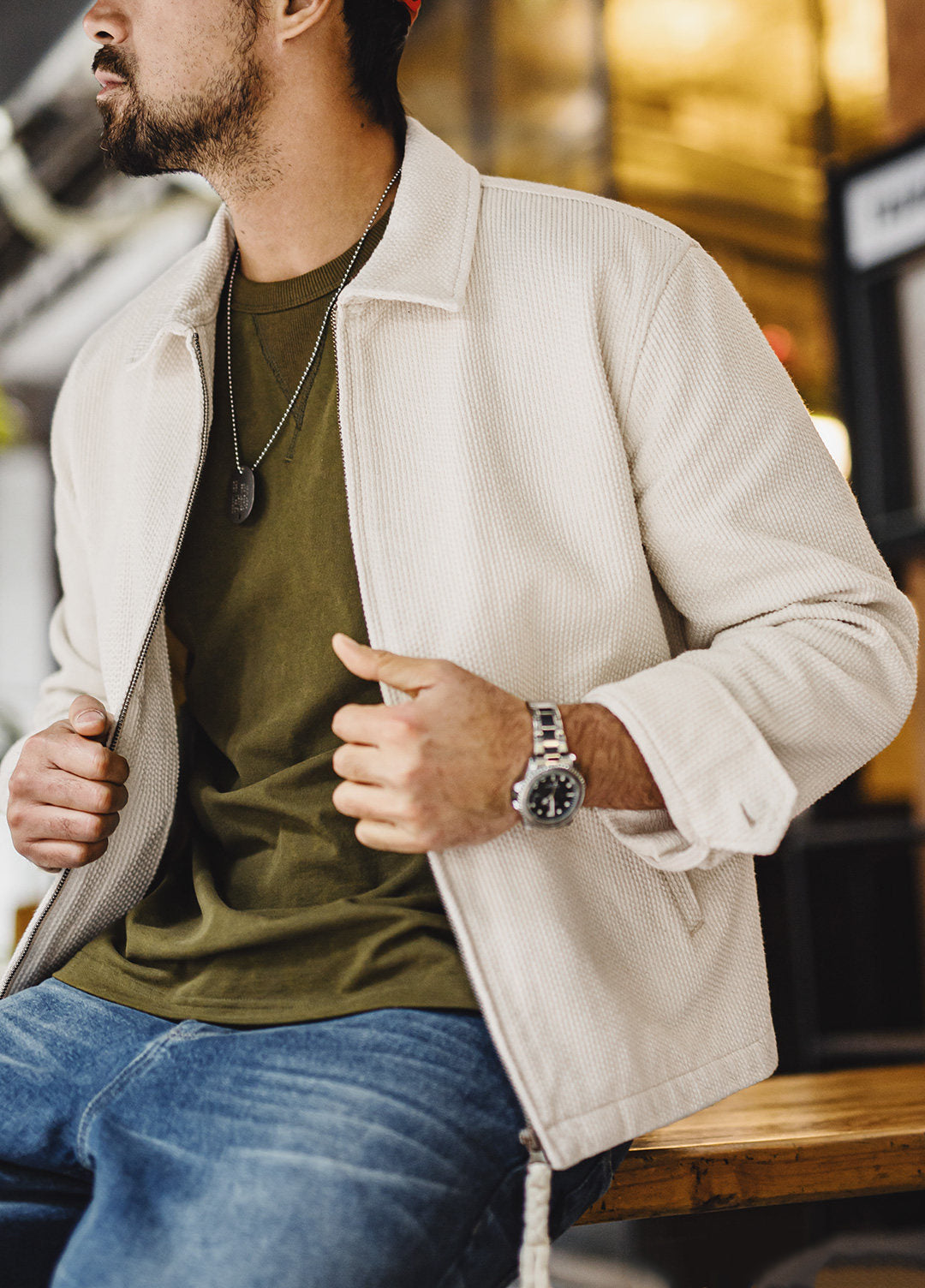 A man wearing a light beige ribbed zip-up jacket over an olive green t-shirt and blue jeans, sitting on a wooden bench in a warm, softly blurred indoor setting.