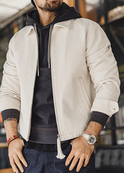 A man wearing a light beige ribbed zip-up jacket over a black hoodie and dark blue jeans, standing in a blurred indoor setting.