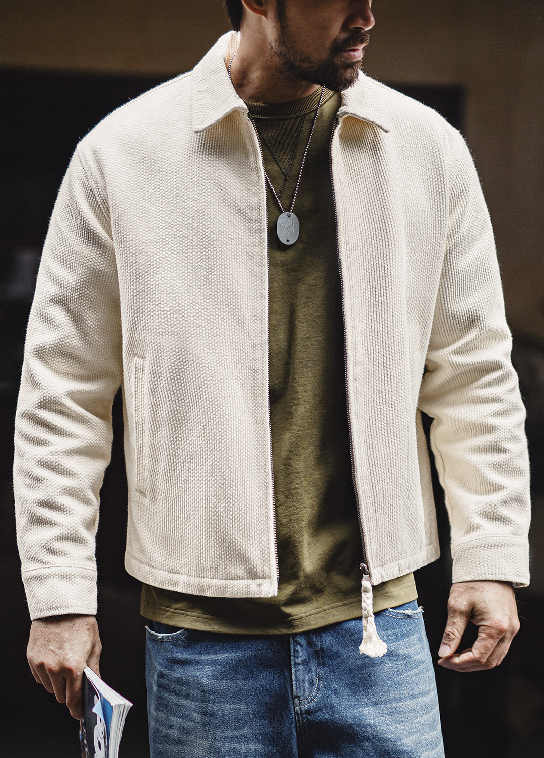 A man wearing a cream-colored textured zip-up jacket over an olive green t-shirt and blue jeans, holding a magazine.