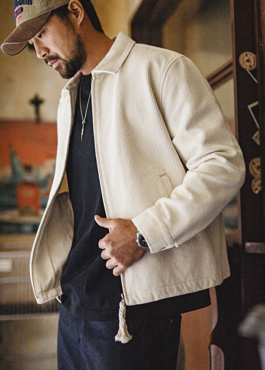 A man wearing a textured off-white corduroy jacket over a black t-shirt and dark jeans, wearing a baseball cap and looking down in an indoor setting.