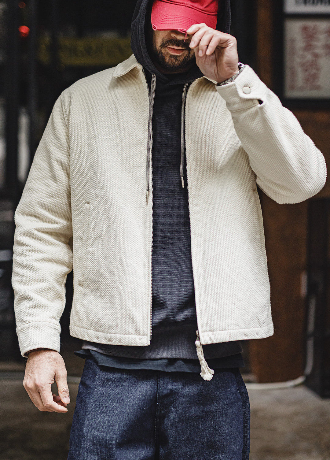 A man wearing a cream-colored textured zip-up jacket over a dark hoodie and blue jeans, with a red cap covering his eyes, standing in an outdoor urban setting.