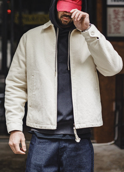 A man wearing a cream-colored textured zip-up jacket over a dark hoodie and blue jeans, with a red cap covering his eyes, standing in an outdoor urban setting.