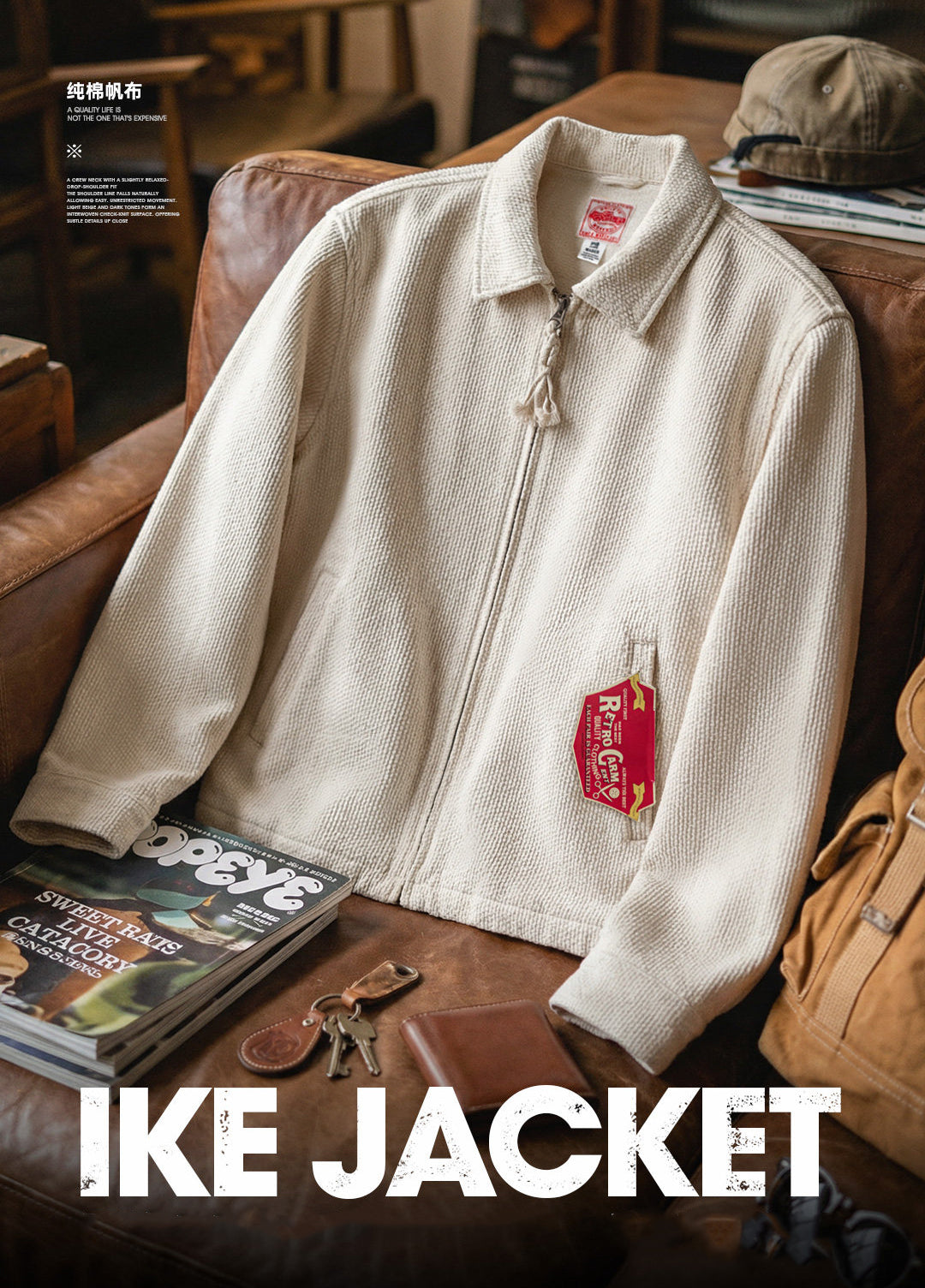 An off-white textured zip-up jacket styled on a brown leather couch with lifestyle props including a cap, keys, a magazine, and a brown bag. The jacket features a collar, a front zipper, and a red and yellow patch on the chest pocket.